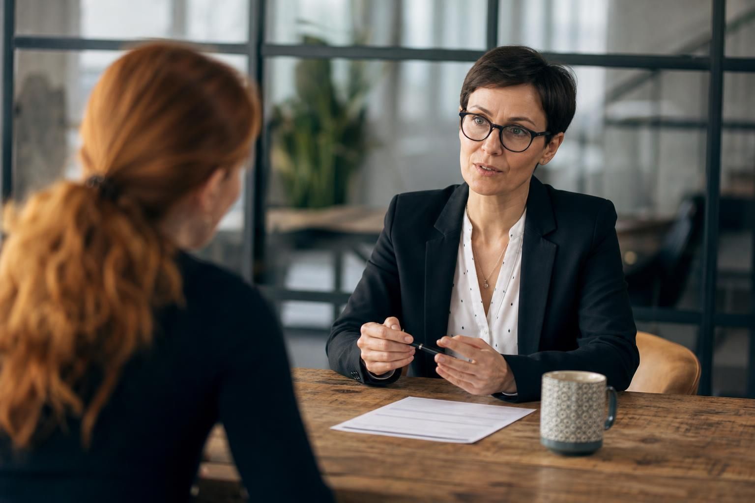 Professional woman in a blazer and glasses conducting an interview with a colleague across a wooden desk in a modern office.